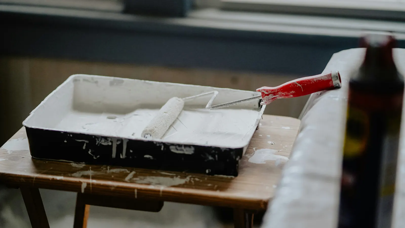 Verfbak met witte verf en een verfroller met rood handvat op een houten krukje bij het raam